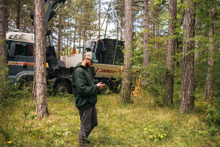 Aufbau im Wald: Ein Kranwagen setzt den Spiegelwürfel ab, vorne ein Crew-Mitglied mit Smartphone